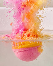 A pink bath bomb dissolving in clear water, creating a dramatic splash of pink, orange, and yellow foam above the surface.