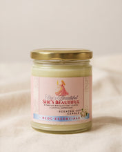 A glass jar candle with a gold metal lid and a pink label reading 'She's Beautiful' by RCDC Essentials, set against a soft beige fabric background.