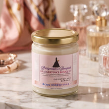 A glass jar candle with a gold lid and a pink label reading Bridgerton's Bouquet by RCDC Essentials, set on a white marble surface with softly blurred perfume bottles and a pink scarf in the background.