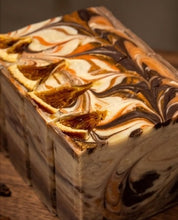 A close-up of a rectangular artisan soap bar with a marbled swirl pattern of dark brown, cream, and amber tones, resting on a wooden surface.