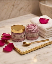 Two jars of whipped soap scrub, one with a gold lid and one open to show the pink scrub inside, styled on a marble tray with a gold spoon, white towels, and pink rose petals.