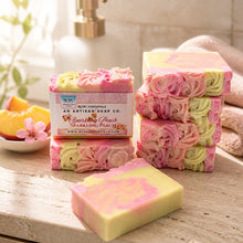 A bar of handmade cold process goat's milk soap with pink and yellow layers, decorated with a floral design, next to a vase with peach flowers.
