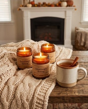 Three amber glass jar candles labeled 'Something's Baking' are arranged on a rustic wooden table draped with a cream cable-knit blanket, alongside a ceramic mug with a cinnamon stick. The background shows a cozy living room setting with a fireplace and autumn decorations.