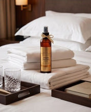 An amber glass room spray bottle with a gold label and ribbon bow, styled on a bed with white linens alongside folded towels, glassware, and wooden trays in a hotel-like setting.