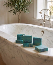 Several turquoise and teal rectangular soap bars with gold glitter details are arranged on the edge of a white marble bathtub in a bright bathroom setting.