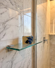 A modern glass wall-mounted shelf with a chrome bracket holding a small blue and yellow soap dish, set against a white marble tiled wall and glass shower door.