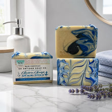 Two artisan soap bars and one wrapped bar from Redc Essentials An Artisan Soap Co. displayed on a marble countertop with a soap dispenser, folded towels, and lavender sprigs in a bright bathroom setting.