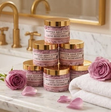 Six jars of whipped soap scrub arranged on a marble bathroom counter with gold faucets, pink roses, and white towels in the background.