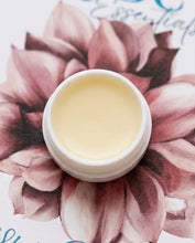 An open white jar of pale yellow balm or perfume balm placed on top of a large pink floral illustration with partial blue script text visible.