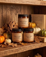Three amber glass jars of Wood Essentials 'Something's Baking' candles arranged on rustic wooden shelves with autumn leaves, pumpkins, and a book.