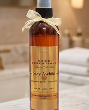 RCDC Essentials Home Collection Stay Awhile room and linen spray in a clear bottle with amber liquid, a black spray top, and a cream ribbon bow, set against a softly blurred bathroom background.