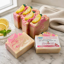 A image of handmade Pink Lemonade soap with pink and yellow colors, decorated with lemon slice designs, displayed with white towel and other decorative items.