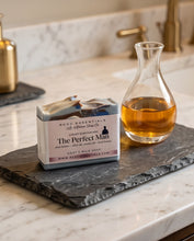 A bar of soap labeled 'The Perfect Man' by BCDC Essentials sits on a dark slate tray beside a glass carafe filled with amber liquid on a marble countertop in a bathroom setting.
