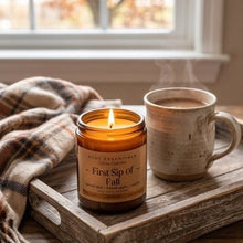 An amber glass jar candle labeled 'First Sip of Fall' by Acme Essentials sits on a rustic wooden tray beside a ceramic mug with steam rising, next to a plaid throw blanket in front of a window.
