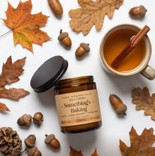 An amber glass jar candle with a black lid and a beige label reading 'Something's Baking' by Herb Essentials, styled on a white background with autumn leaves, acorns, a mug of tea with a cinnamon stick, and text at the bottom that says '4/4 GRAB YOUR COZY VIBES'.