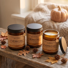 Three amber glass scented candles with black lids from the brand ROCC ESSENTIALS, labeled 'Fall Into Cozy', styled on a rustic wooden table with autumn leaves, pinecones, and a cozy knitted blanket in the background.