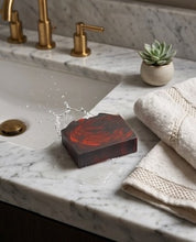 A rectangular bar of soap with a dark reddish-brown marbled pattern sits in a white sink on a white marble countertop. A gold faucet and handle are visible behind it, along with folded cream towels and a small potted succulent.