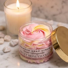 A jar of whipped soap scrub labeled 'Sweet Honeysuckle' with pink and yellow swirled soap inside, shown on a marble surface with a gold lid beside it and a lit candle in the background.
