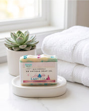 A bar of handmade soap from Lady Lola Artisan Soap Co. sits on a white dish in a bright bathroom setting, with a small succulent and folded white towels in the background.