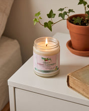 A lit candle in a clear glass jar sits on a white side table in a cozy indoor setting, with a potted ivy plant and an old book in the background.