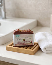 A bar of soap from An Artisan Soap Co. sits on a wooden soap dish on a bathroom counter next to a folded white towel.