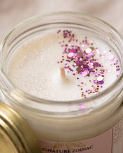 A close-up of a glass jar candle with white wax, a wooden wick, and scattered pink, purple, and metallic glitter on top. Part of the gold-toned lid is visible at the lower left, and a pink label reads 'SIGNATURE ZODIAC'.