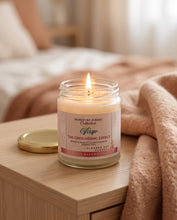 A lit candle in a clear glass jar sits on a light wood bedside table next to a beige blanket in a softly blurred bedroom setting. The label reads 'Signature Zodiac Collection' and 'Wingo The Grounding Effect'.