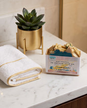 An open box of artisan soap on a white marble countertop, next to a folded white towel with gold trim and a small potted succulent in a gold planter. The soap is labeled 'Ocean Ember' by 'BCBC Essentials' and 'An Artisan Essentials Soap Co.