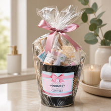 Gift set with a candle and bath products, wrapped in cellophane with a pink ribbon, on a kitchen counter.