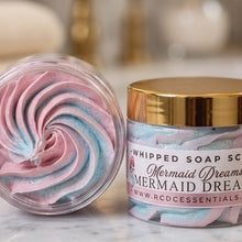 A jar of whipped soap scrub labeled 'Mermaid Dreams' by RCDC Essentials, shown next to an open clear lid revealing pink and blue swirled whipped soap inside, set on a marble surface with a blurred bathroom background.