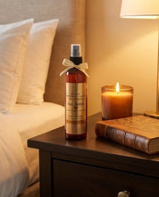 A bottle of Leo G. Essentials 'Sex Aesthetic' body spray stands on a dark wood bedside table next to a lit candle and a vintage book, with a softly lit bed in the background.