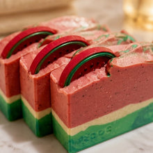 Three rectangular layered soap bars with a watermelon design, featuring a pink top layer, green middle layer, cream bottom layer, and a red watermelon slice garnish on top, set on a light marble surface.