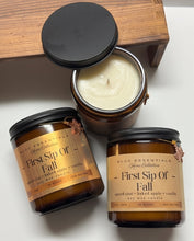 Three amber glass soy wax candles with black lids are arranged on a white surface with a wooden block. One candle is open, showing the cream-colored wax, while the other two have labels reading 'First Sip Of Fall' and 'SCC Essentials Home Collection.