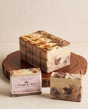 Three artisanal salt and balm soap bars with a marbled swirl pattern in brown, beige, and gold tones, displayed on a rustic wooden slice stand against a textured off-white wall.