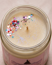 Top-down view of a glass jar candle filled with creamy beige wax, decorated with colorful round glitter confetti and fine silver holographic glitter. The pink label at the bottom reads 'Bridgero's Boutique'.