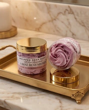 A jar of whipped soap scrub and a matching rose-shaped bath bomb on a gold tray, set on a marble countertop with a candle in the background.