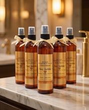 Five amber glass room linen sprays with gold labels and black spray tops are lined up on a marble countertop in a warm, softly lit bathroom setting.
