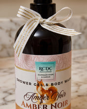 A bottle of RCDC Essentials Amber Noir Shower Gel & Body Wash, featuring a dark amber glass bottle, a pink and white label with a fox illustration, and a cream ribbon with gold stripes tied in a bow around the neck.