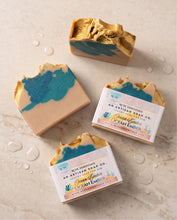 Four artisan soap bars from The HCK Emporium, specifically Ocean Ember, arranged on a light beige marble surface with decorative water droplets. Three bars are wrapped in white paper packaging, and one is unwrapped to show its marbled texture of teal blue and golden amber soap layers.