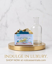 A bar of artisan soap in a small box sits on a round gold tray beside a clear glass bottle, set against a white marble background. Text at the bottom reads 'INDULGE IN LUXURY.