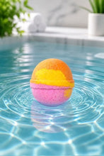 A colorful spherical bath bomb with orange, yellow, and pink layers floats in the center of a swimming pool, creating ripples on the clear blue water.