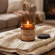 An amber glass jar candle labeled 'Fall Into Cozy' by SCDC Essentials sits lit on a wooden table draped with a cream cable-knit blanket, with its black lid placed beside it. The background shows a softly blurred living room with a sofa and fireplace.