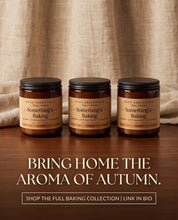 Three amber glass jars with black lids and tan labels reading 'Something's Baking' are arranged on a wooden surface in front of a beige fabric backdrop. Large text below says 'BRING HOME THE AROMA OF AUTUMN.