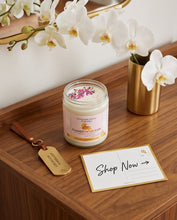 A jar candle labeled Summer Nights Scandi sits on a wooden table beside a gold key tag and a 'Shop Now' postcard, with white orchids and a gold vase in the background.