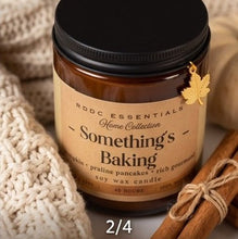 An amber glass soy wax candle jar with a black lid and a tan label reading 'Something's Baking' and 'RDDC Essentials Home Collection,' styled with a knitted cream textile, cinnamon sticks, and a small gold leaf charm. Text overlay at the bottom reads '2/4 PUMPKIN PRALINE PERFECTION.