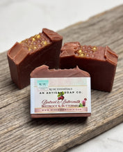 Three handmade soap bars with a rustic finish and gold honeycomb details, arranged on a wooden board. The center bar has a label for 'Red DC Essentials' 'Beetroot & Buttermilk' soap.
