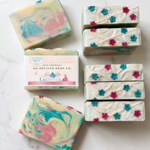 Six artisan soap bars and one wrapped bar from Lady Ola Botanical Essentials arranged on a white marble surface. The soaps feature marbled swirls of pastel pink, blue, and green, as well as white bars decorated with small pink and blue flower motifs.