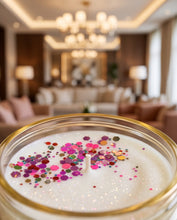 A close-up of a white candle in a clear glass jar with a gold rim, decorated with colorful glitter and confetti, set against a blurred modern living room background.