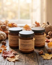 Three amber glass jars of scented candles labeled 'Fall Into Cozy' by Coc Essentials, arranged on a rustic wooden table with autumn leaves, acorns, pumpkins, and a softly blurred window background.