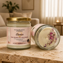 Two glass scented soy candles from the Signature Zodiac Collection, one standing upright and one lying on its side, displayed on a stone surface with a softly blurred living room background.